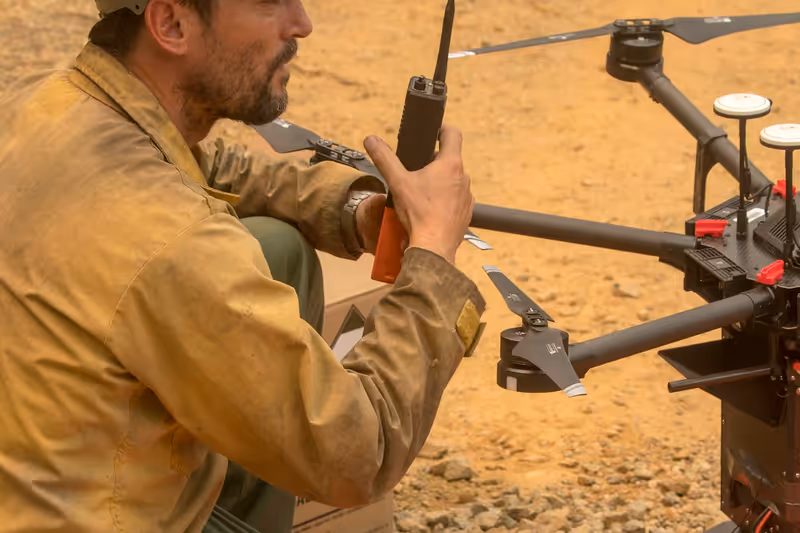 Combatiente forestal con camisa ignífuga y casco prepara un dron sobre su plataforma mientras usa el radio