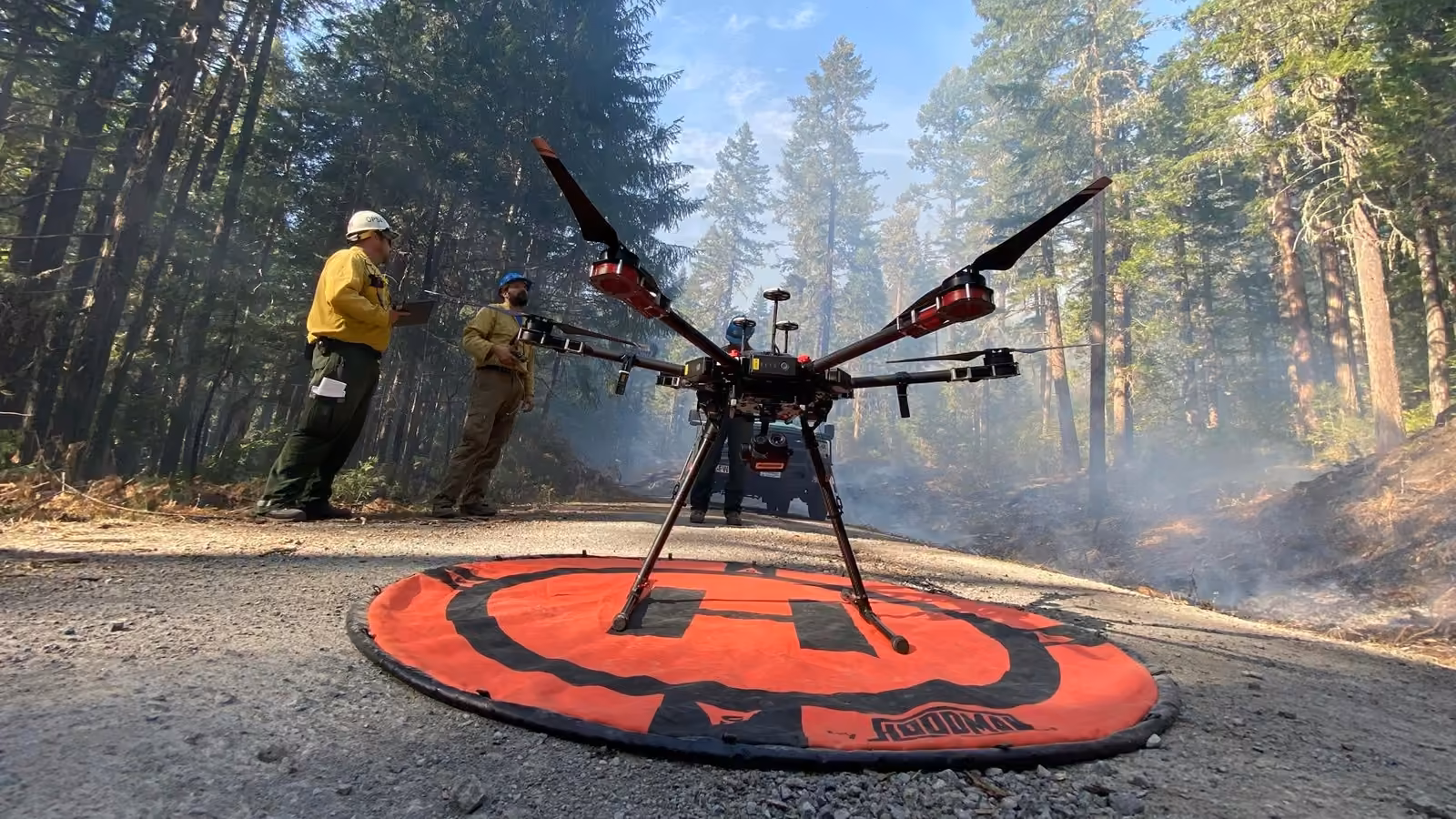 Dron hexacóptero sobre su plataforma de despegue en un camino forestal con humo, mientras dos combatientes de incendios preparan el vuelo de reconocimiento