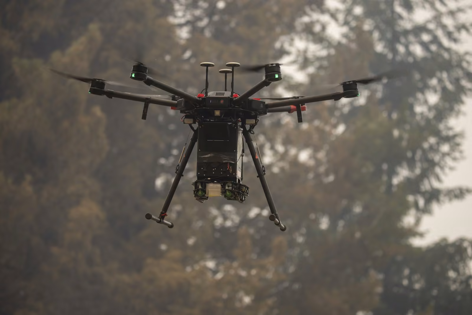 Dron hexacóptero con cámara y carga útil en vuelo frente a un bosque cubierto de humo durante un incendio forestal