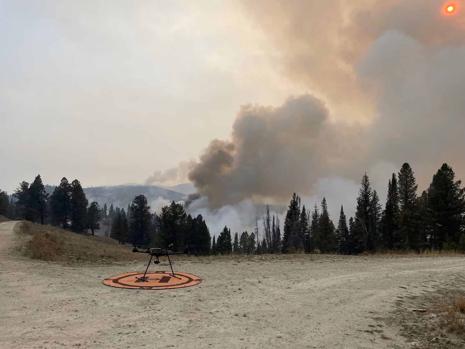 Dron listo para despegar desde una plataforma en un claro, frente a una columna de humo de un incendio forestal en zona de pinos