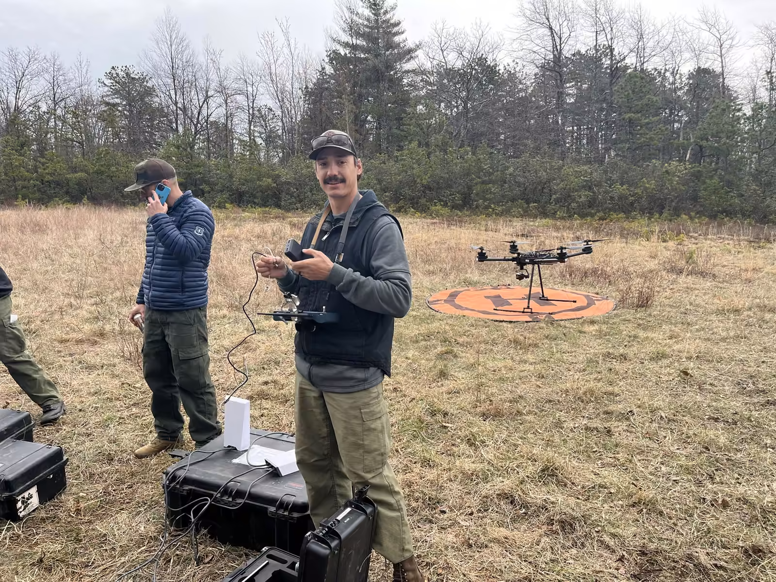 Pilotos con control remoto, estuches de transporte y un dron sobre su plataforma en campo abierto