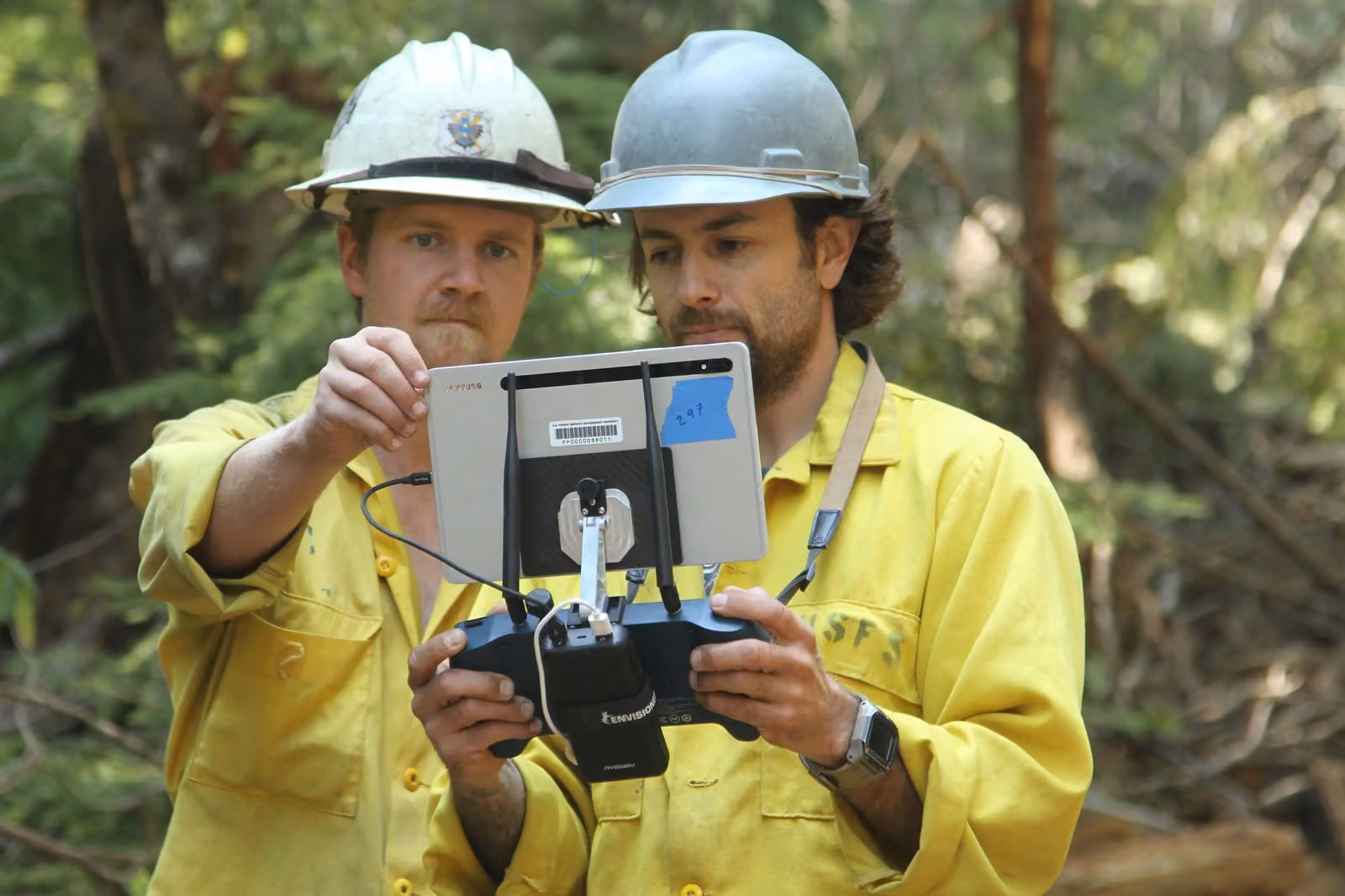 Dos combatientes de incendios forestales con casco revisan en la pantalla del control remoto el video transmitido por un dron